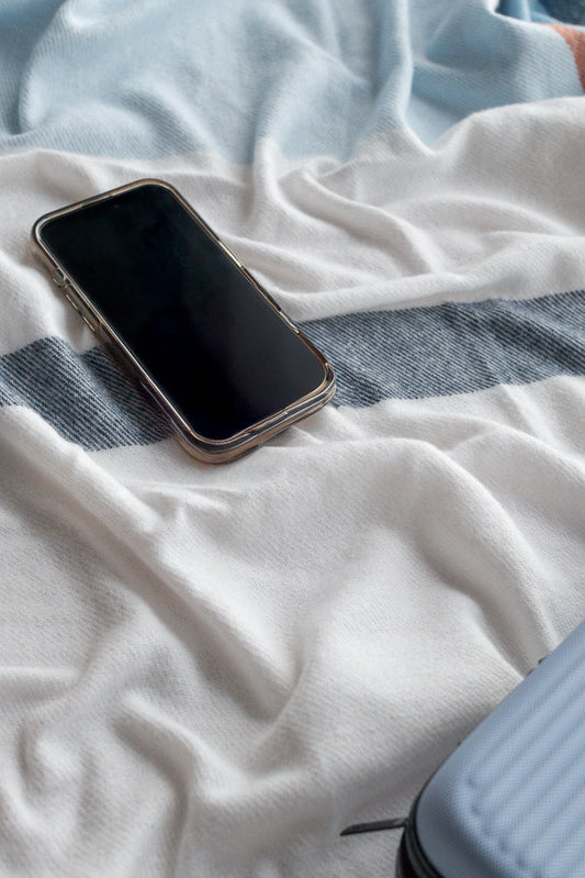 A smartphone with a black screen rests on a white and blue striped blanket. The blanket is wrinkled, creating soft folds and shadows around the phone.