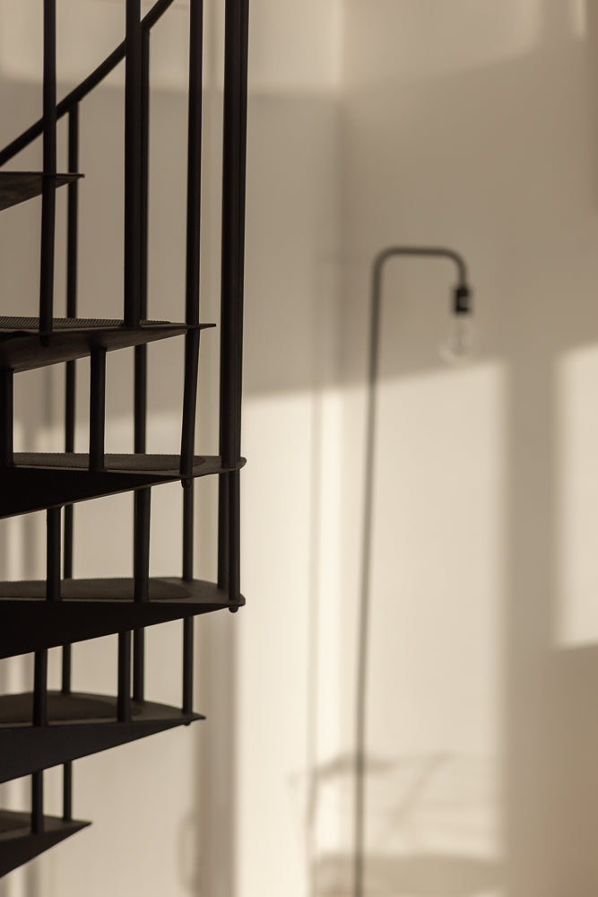 A close-up, low-angle shot of a black spiral staircase against a light-colored wall. The staircase is in focus on the left side of the frame, with its metal steps and railings clearly visible. In the background, a floor lamp is out of focus, casting a shadow on the wall.