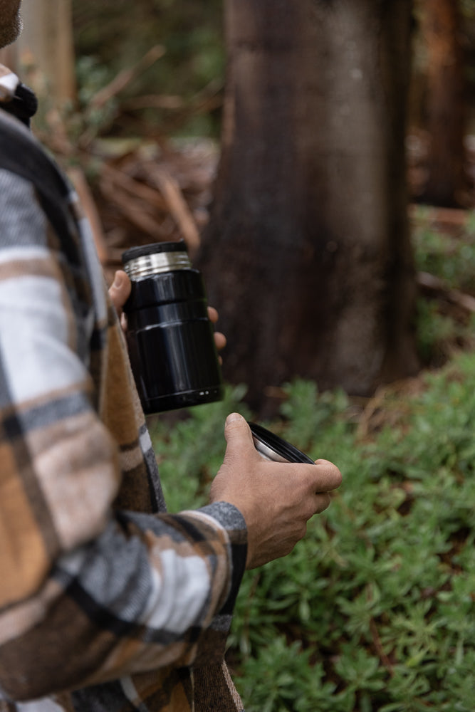A person wearing a plaid jacket holds a black thermos and its lid in a forest setting.