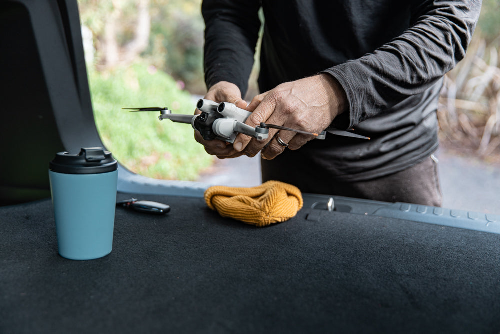 A person in a black long-sleeved shirt holds a small white drone with black propellers. A blue travel mug and a yellow knitted hat sit on a dark surface next to the drone.