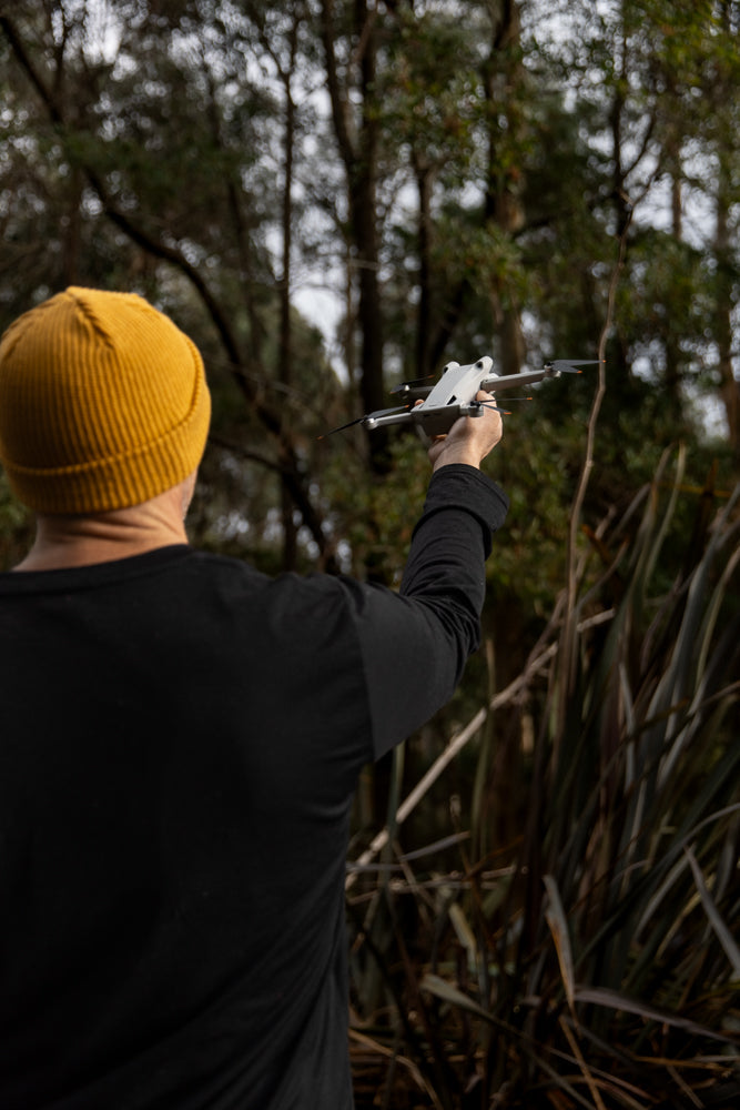 A person wearing a yellow beanie and black shirt holds a white drone in their outstretched hand, ready for flight amidst trees and foliage.