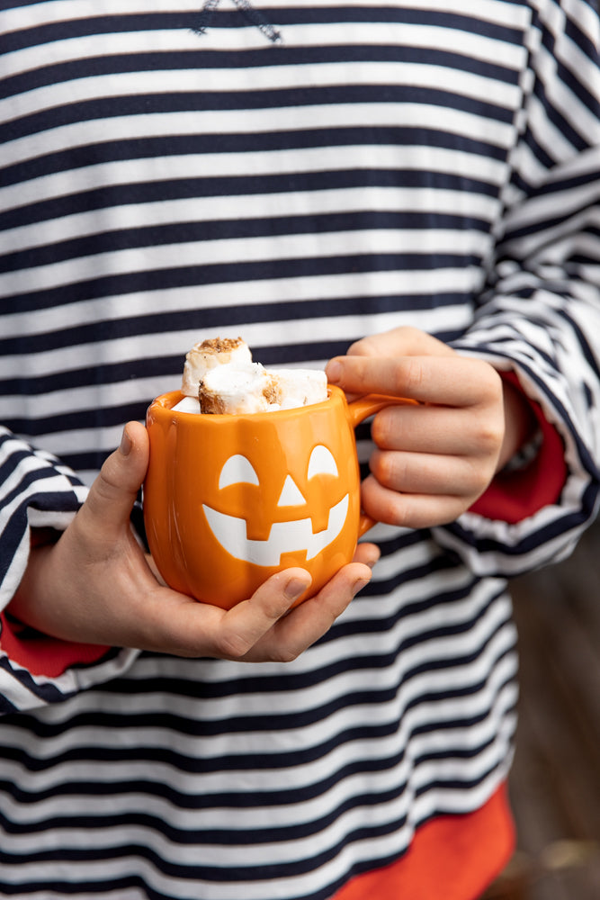 A child in a striped shirt holds a pumpkin-shaped mug filled with marshmallows.