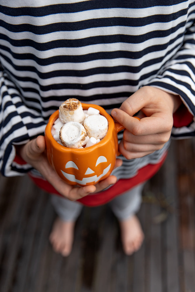 A child wearing a striped shirt holds a small orange pumpkin-shaped mug filled with marshmallows and toasted marshmallows.
