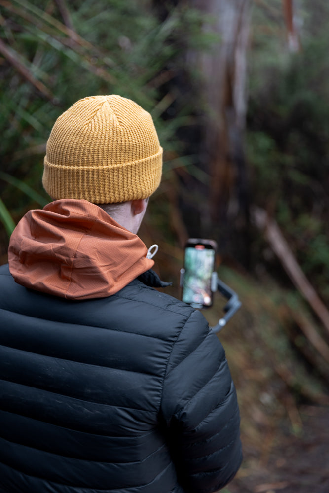 A person wearing a yellow beanie and a black puffer jacket with an orange hood is filming with a smartphone mounted on a gimbal in a forest.