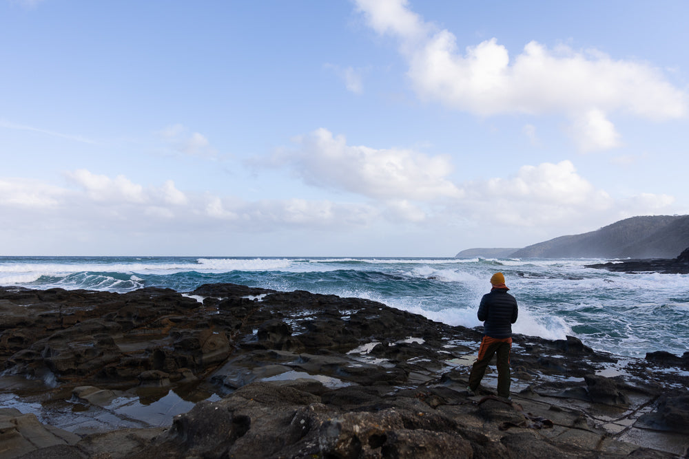 A person wearing a yellow hat and a puffer jacket stands on dark, rocky shore, looking out at the choppy, blue ocean under a cloudy sky.