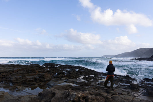 A person wearing a yellow hat and a puffer jacket stands on dark, rocky shore, looking out at the choppy, blue ocean under a cloudy sky.