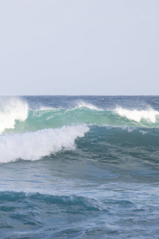 A series of ocean waves roll towards the shore under a pale blue sky. The waves are a mix of deep blue and turquoise, with white foam cresting on the larger waves.