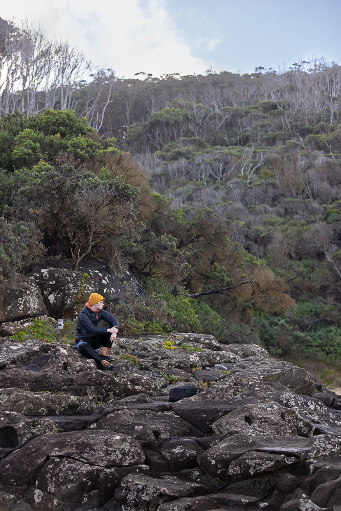 A teenage boy sitting on a rock cliff looking out surrounded by trees and hills