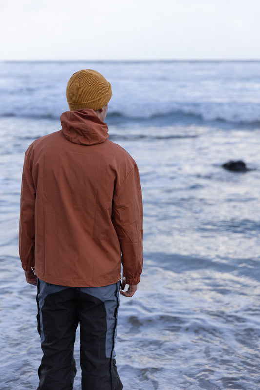 A person wearing a rust-colored jacket and a yellow beanie stands on a beach, looking out at the ocean waves.