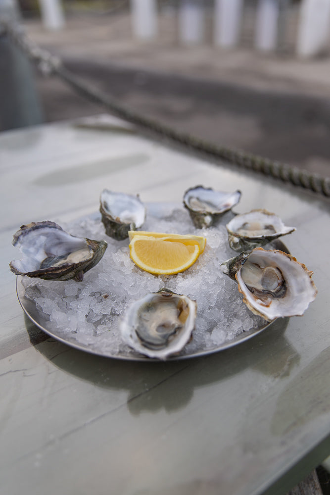 A plate of six fresh oysters on a bed of crushed ice, garnished with a slice of lemon. The oysters are served in their shells and appear plump and juicy.