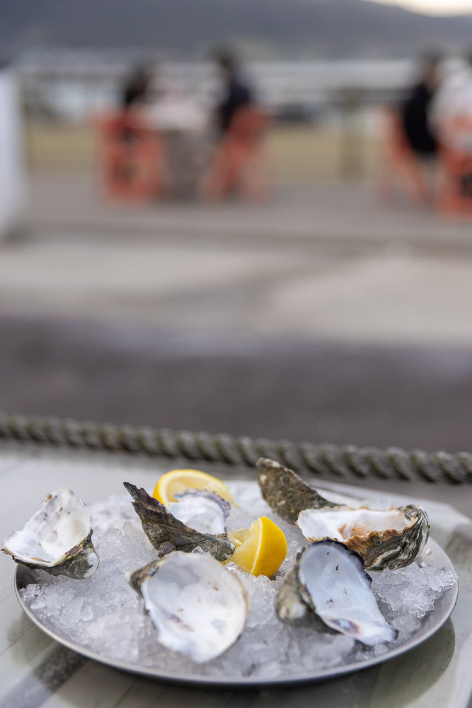 A platter of fresh oysters on ice with lemon wedges. In the background, blurred figures are seated at tables outdoors.