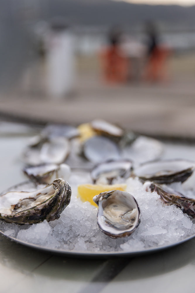 A close-up shot of a platter of fresh oysters on crushed ice, garnished with a lemon wedge. The oysters are partially open, revealing their glistening flesh.