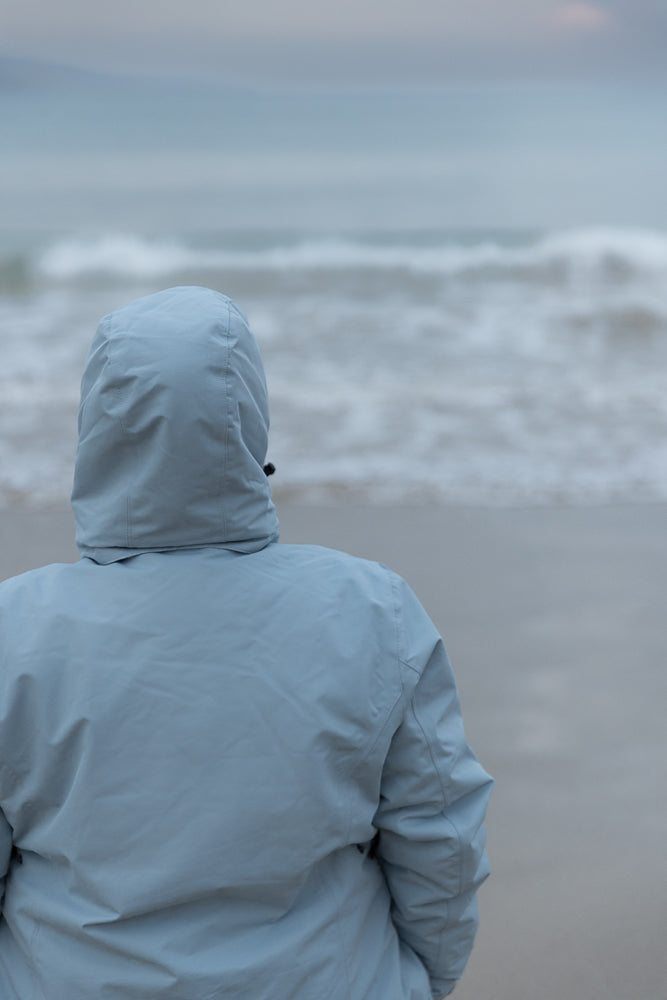 A person wearing a light blue hooded jacket stands on a beach, facing away from the camera towards the ocean. The sky is overcast and the waves are choppy.