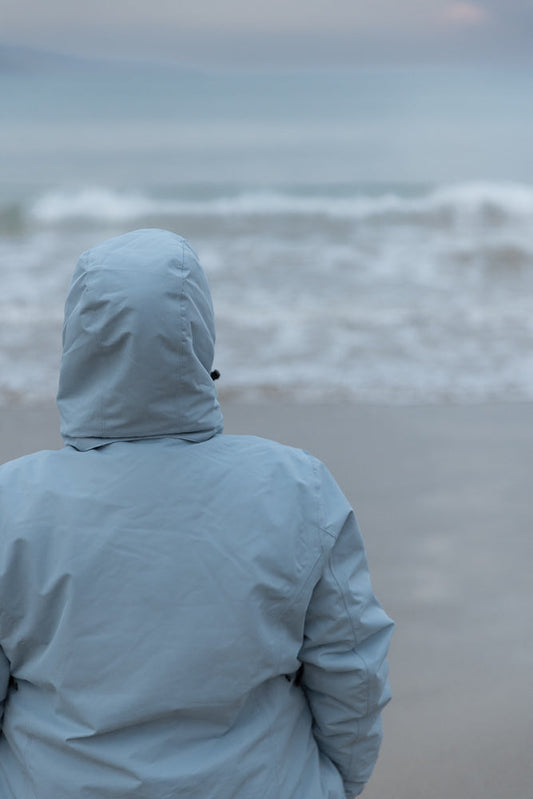 A person wearing a light blue hooded jacket stands on a beach, facing away from the camera towards the ocean. The sky is overcast and the waves are choppy.
