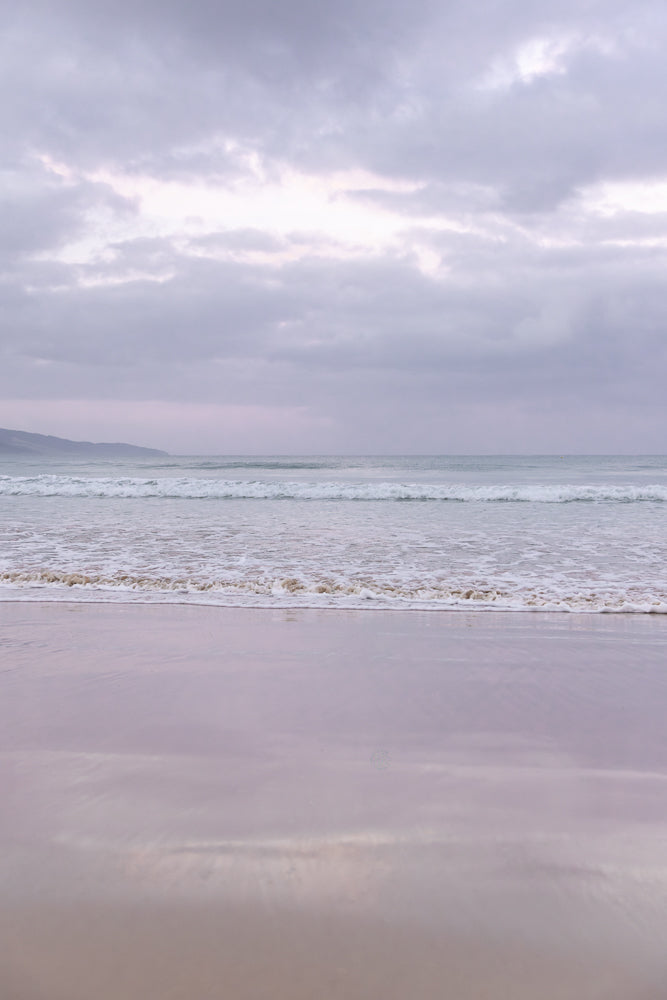 A serene beach scene with gentle waves rolling onto the shore under a cloudy, overcast sky. The sand reflects the soft light, creating a peaceful and muted atmosphere.