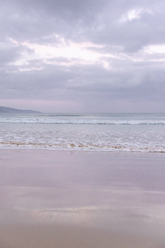 A serene beach scene with gentle waves rolling onto the shore under a cloudy, overcast sky. The sand reflects the soft light, creating a peaceful and muted atmosphere.