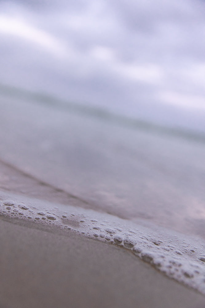 A close-up, low-angle shot of gentle waves lapping onto a sandy shore. The water is calm and creates a thin line of foam and bubbles along the edge of the sand. The background is blurred, showing a soft, overcast sky.