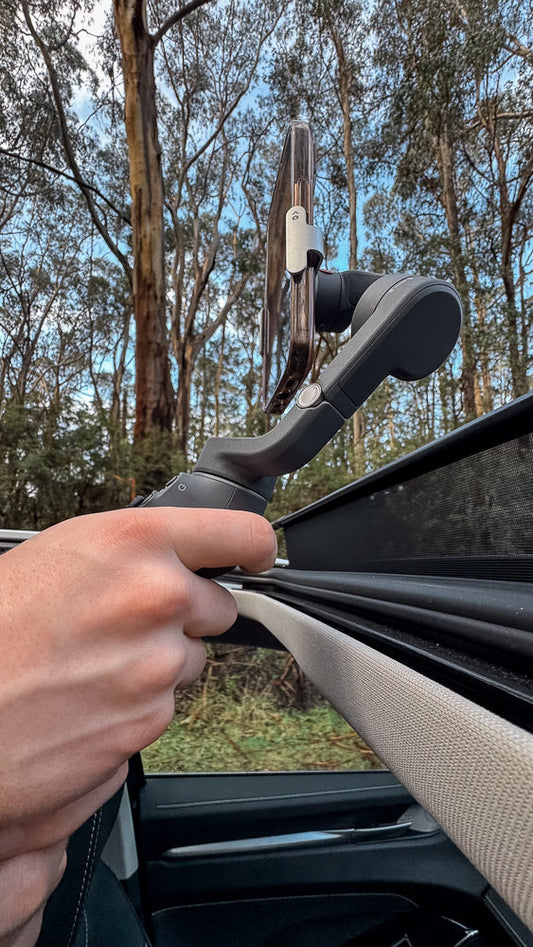 A hand holds a smartphone mounted on a gimbal stabilizer, with the sunroof open and trees visible in the background.