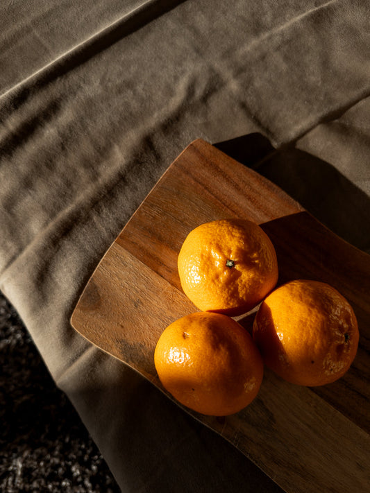 Three bright orange tangerines sit on a wooden cutting board. The board is placed on a textured, taupe-colored fabric. Sunlight casts a diagonal shadow across the scene, highlighting the bumpy texture of the fruit.