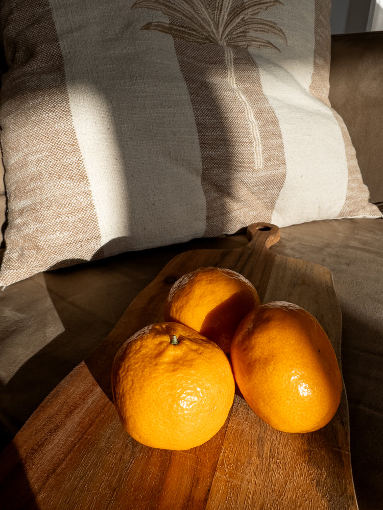 Three bright orange tangerines sit on a wooden cutting board. A striped pillow with a palm leaf design is in the background, with sunlight casting shadows across the scene.