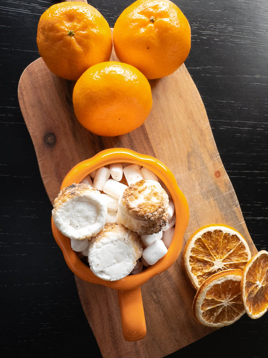 A top-down view of an orange mug filled with marshmallows and toasted marshmallows, surrounded by whole tangerines and dried orange slices on a wooden board.