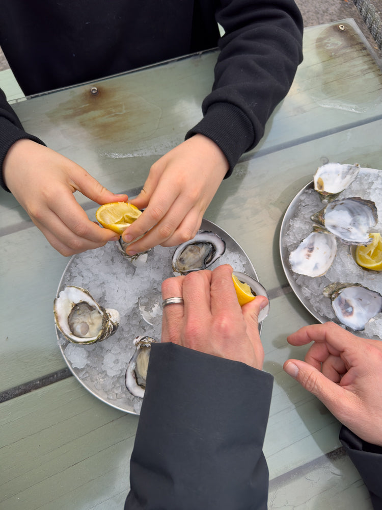 Two people are preparing to eat oysters on a table. One person is squeezing a lemon wedge over a plate of oysters on ice, while another person holds a lemon wedge over their own plate.