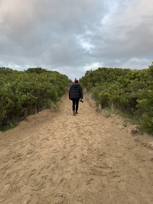 A person wearing a black puffer jacket and a red and white beanie walks away from the camera down a sandy path lined with green bushes under a cloudy sky. The person is holding a camera in their right hand.