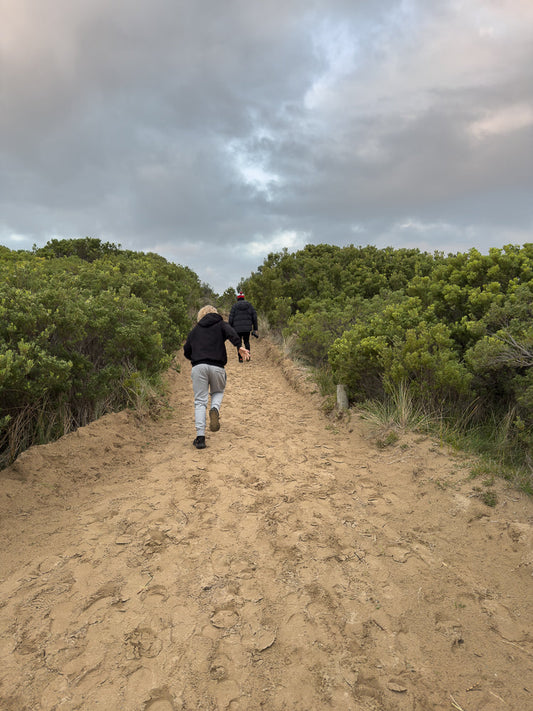 Two people walk up a sandy path lined with green bushes under a cloudy sky. The person in front is wearing a black hoodie and grey pants, while the person behind them wears a black puffer jacket and a red and white hat.