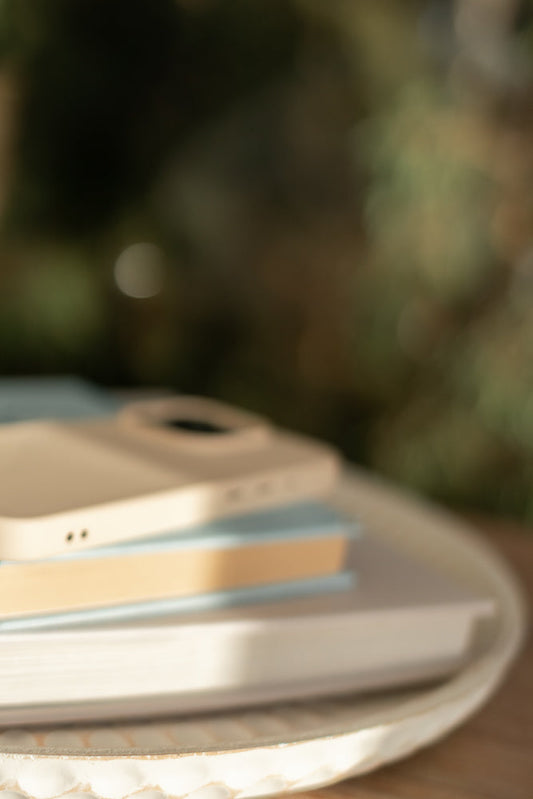 A close-up, slightly blurred shot shows a stack of books on a decorative tray. A light-colored phone case rests on top of the books, with the camera lens visible.