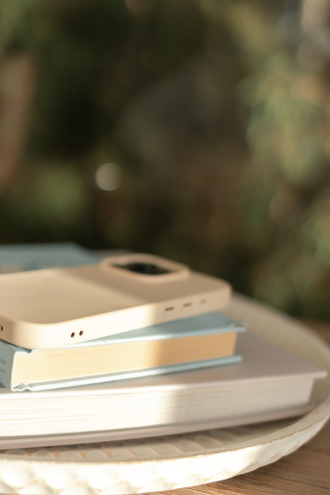 A light beige phone case rests on top of two books, one blue and one cream-colored, arranged on a textured white tray. Sunlight streams across the scene, highlighting the objects against a blurred green background.