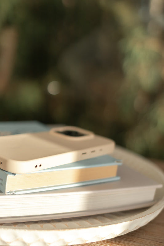 A light beige phone case rests on top of two books, one blue and one cream-colored, arranged on a textured white tray. Sunlight streams across the scene, highlighting the objects against a blurred green background.