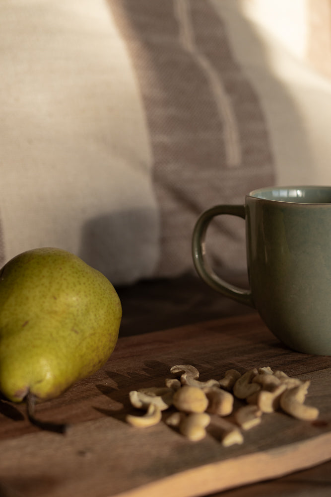 A green pear and a pile of cashews sit on a wooden cutting board. A teal mug is to the right of the cashews. A striped blanket is in the background.