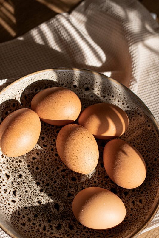 Six brown eggs are arranged in a speckled brown and cream ceramic bowl. Sunlight casts striped shadows across the eggs and the bowl.