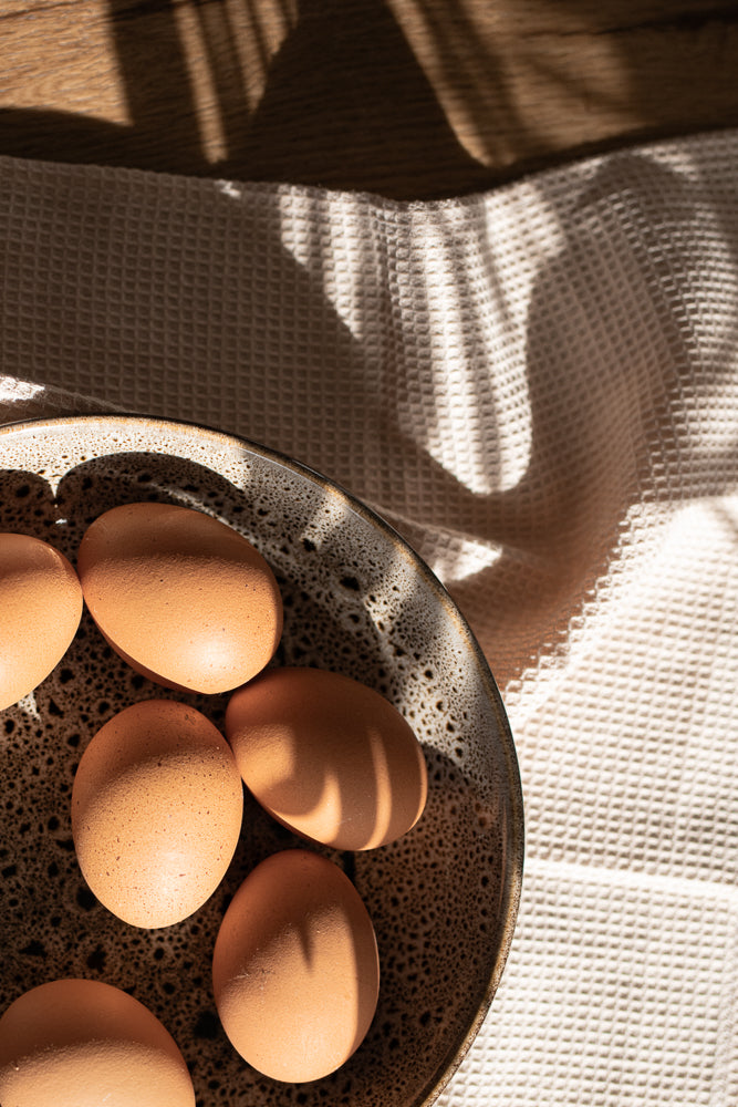 A close-up shot of several brown eggs in a speckled ceramic bowl, with dappled sunlight casting shadows on a textured white cloth in the background.