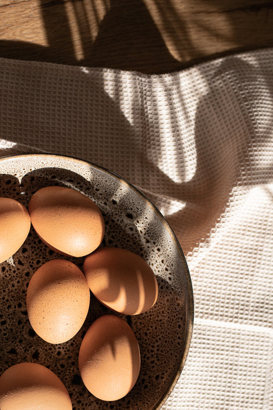 A close-up shot of several brown eggs in a speckled ceramic bowl, with dappled sunlight casting shadows on a textured white cloth in the background.