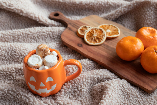 A Halloween-themed orange mug shaped like a jack-o'-lantern is filled with marshmallows and sits on a fluffy cream blanket. Beside it on a wooden cutting board are several clementines and dried orange slices.