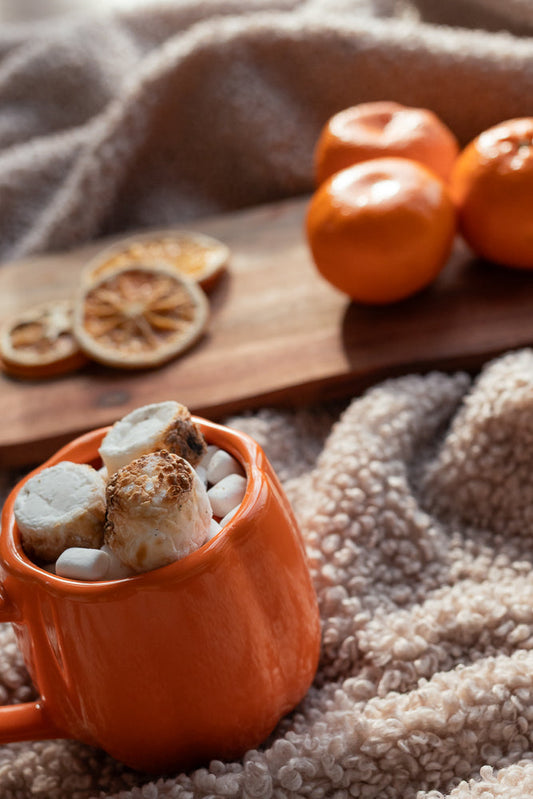 An orange mug filled with toasted marshmallows and small white candies sits on a fluffy cream blanket. In the background, a wooden board holds several clementines and dried orange slices.