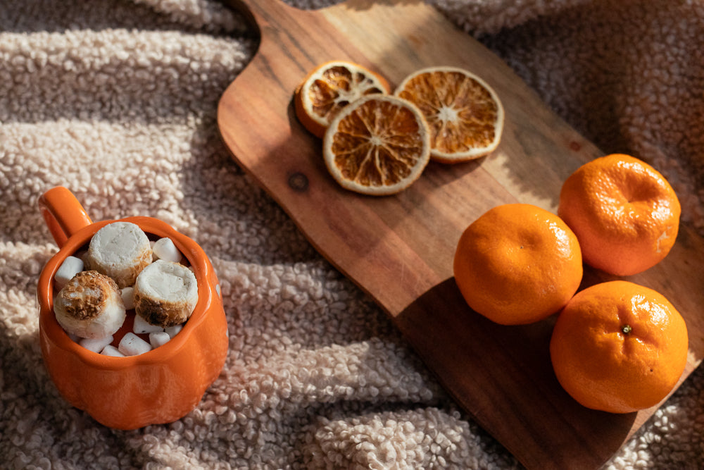 An orange mug filled with marshmallows and toasted marshmallows sits on a fuzzy blanket next to a wooden cutting board with tangerines and dried orange slices.