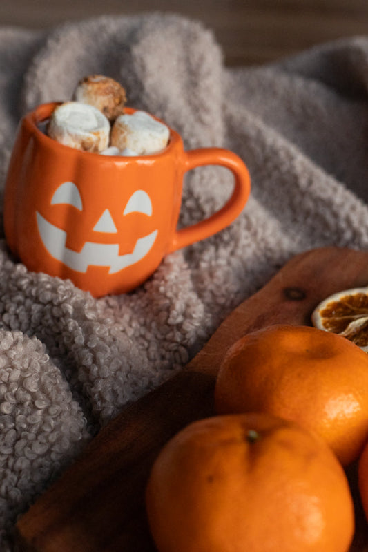 A Halloween-themed orange mug shaped like a jack-o'-lantern is filled with marshmallows and sits on a soft, fuzzy blanket. In the foreground, two clementines and a dried orange slice rest on a wooden board.