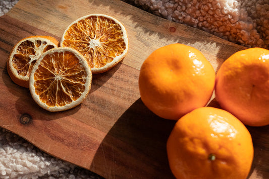 Three whole tangerines and three dried orange slices are arranged on a wooden cutting board. The tangerines are bright orange and glossy, while the orange slices are translucent and show their segmented interior.