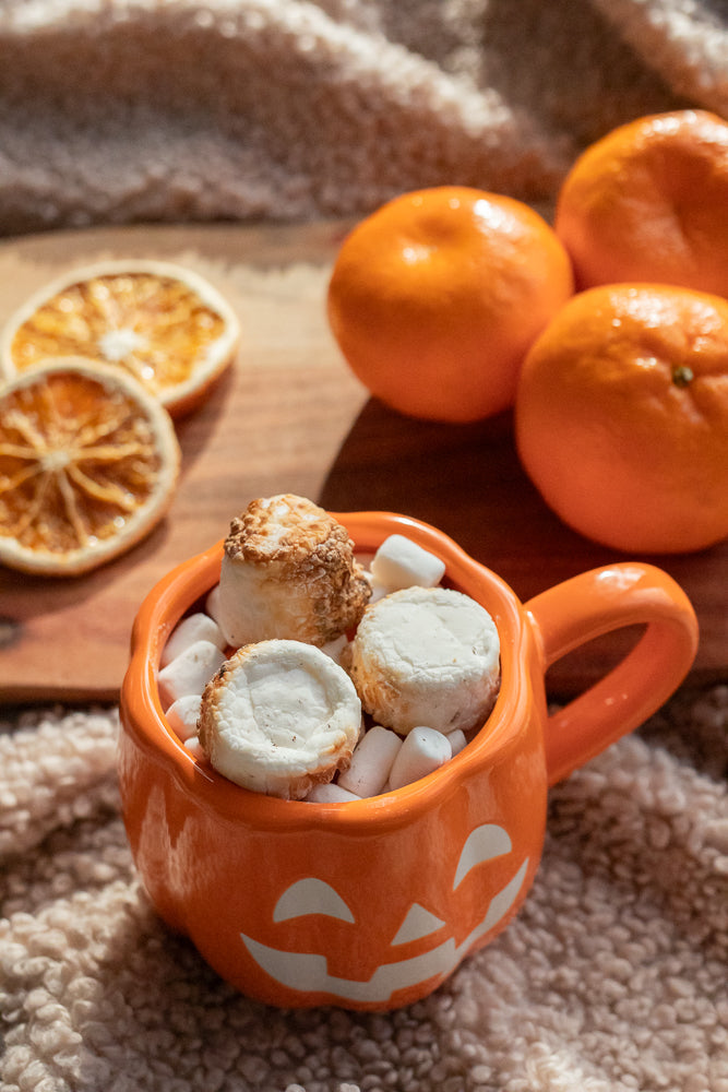 A Halloween-themed orange mug shaped like a jack-o'-lantern is filled with marshmallows and topped with toasted marshmallows. Several whole tangerines and dried orange slices are visible in the background.