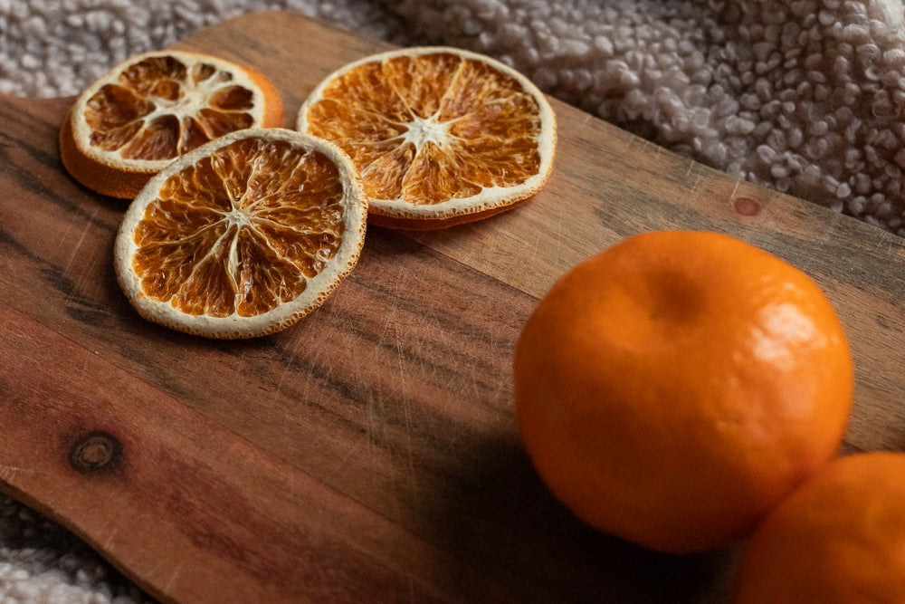 Three dried orange slices and two whole oranges are arranged on a wooden cutting board. The dried slices are on the left, showing their intricate patterns. The whole oranges are on the right, with one in focus and another partially visible.