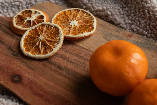 Three dried orange slices and two whole oranges are arranged on a wooden cutting board. The dried slices are on the left, showing their intricate patterns. The whole oranges are on the right, with one in focus and another partially visible.