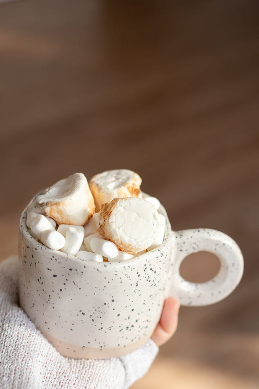 A hand wearing a white knitted sleeve holds a speckled mug filled with mini marshmallows and topped with larger, toasted marshmallows. The mug is white with black speckles and has a handle. The background is a blurred wooden surface.