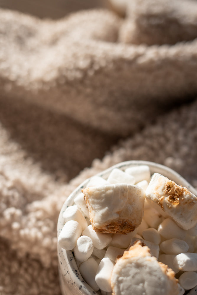 A close-up shot of a bowl filled with marshmallows. Some marshmallows are toasted and golden brown, while others are white and fluffy. The bowl is placed on a soft, textured, beige blanket.