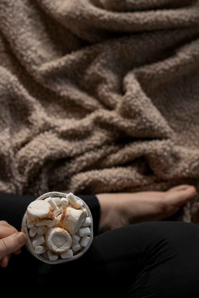 A person's hand holds a mug filled with marshmallows. The person is wrapped in a fuzzy brown blanket, with their bare foot visible in the background.