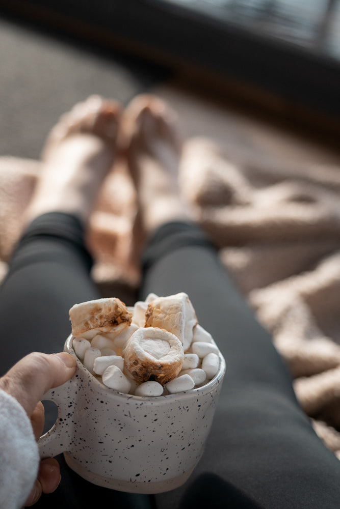 A person holds a mug filled with hot chocolate and marshmallows, with their feet visible in the background, wrapped in a blanket.