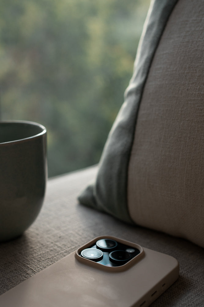 A close-up shot of a smartphone with a triple-lens camera system, resting on a textured surface next to a mug and a pillow. The background is softly blurred, showing greenery outside a window.