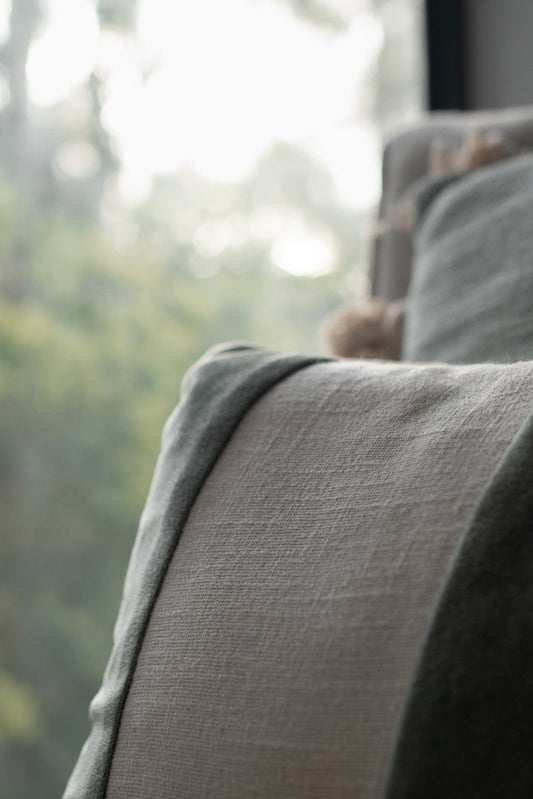 Close-up of a textured light beige fabric armchair with a dark green throw blanket draped over the armrest. The background is softly blurred, showing a bright, out-of-focus window with greenery.