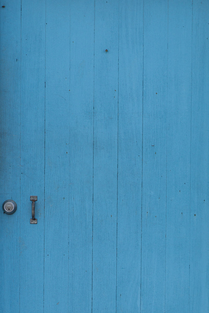 A close-up, vertical shot of a weathered, bright blue wooden door with vertical planks. A silver doorknob and lock are visible on the left side of the door.